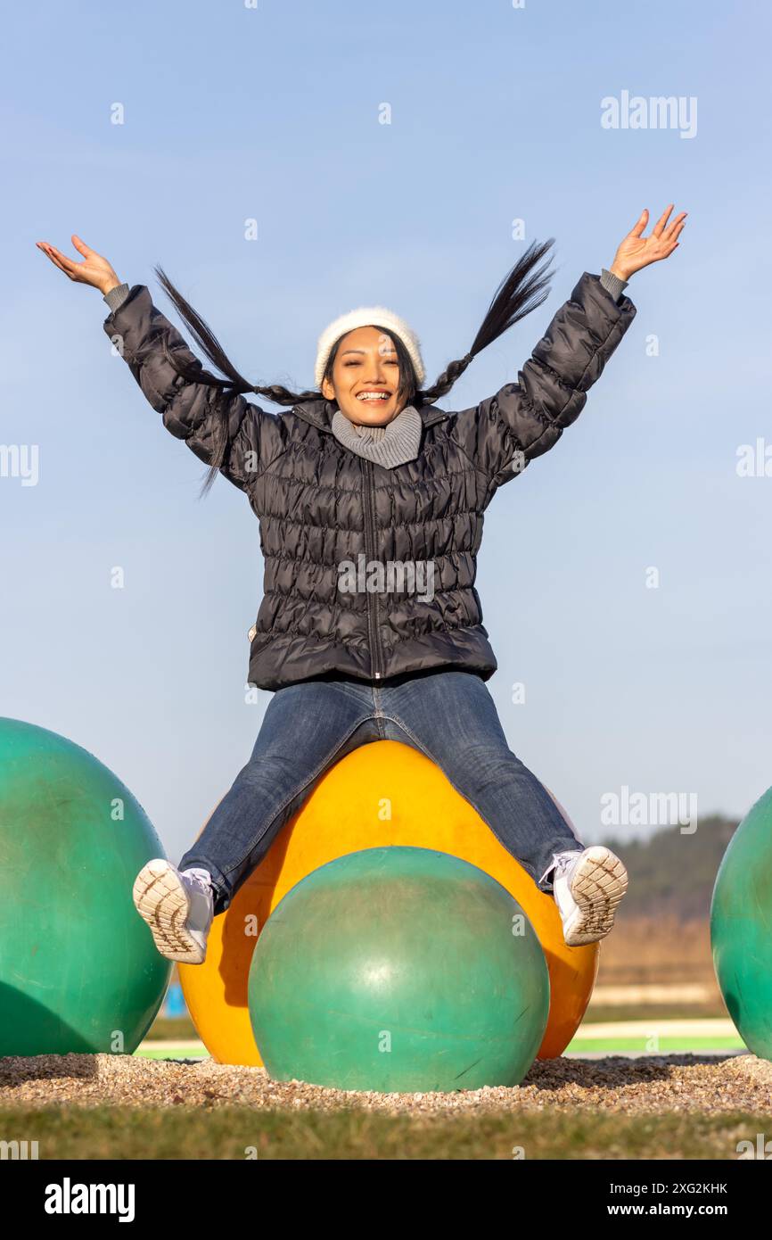 A cheerful woman is sitting on a large colorful ball on the playground Stock Photo