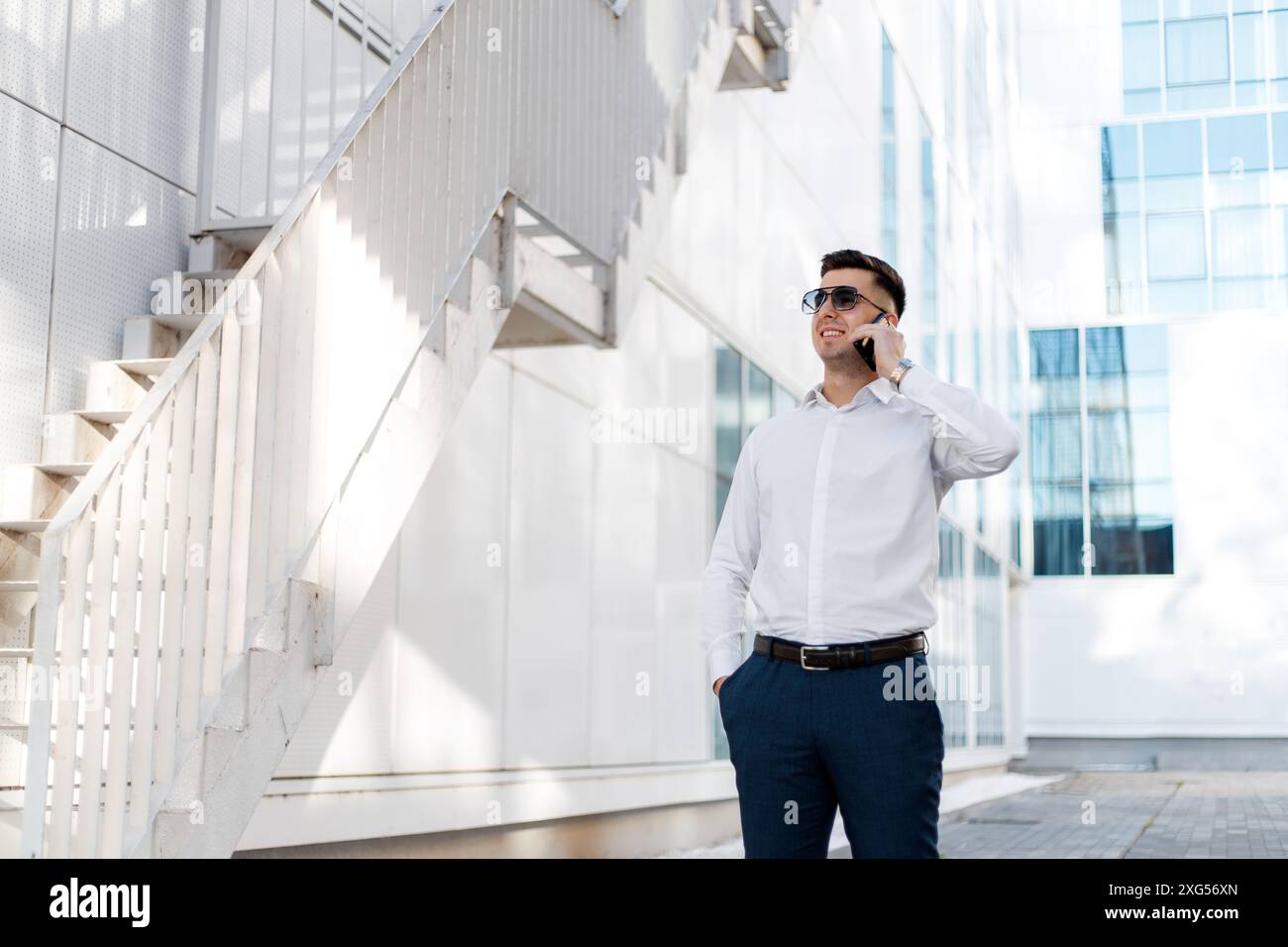 A middle-aged man is standing in front of a modern office building, engaged in a phone call on his mobile device. Stock Photo