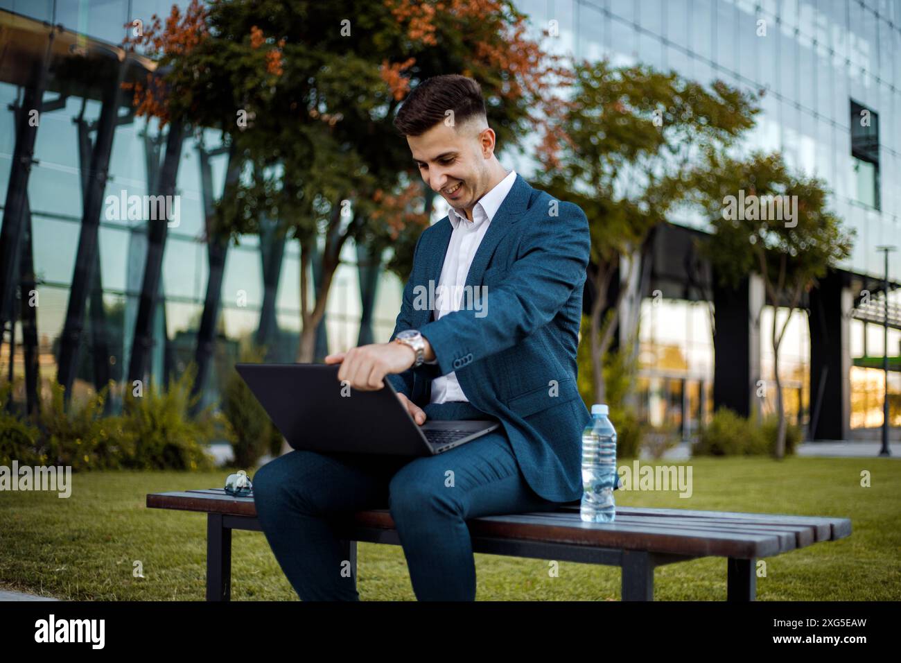 A man dressed in a formal suit is sitting on a bench outdoors. He is focused on his laptop screen, possibly working or studying. Stock Photo