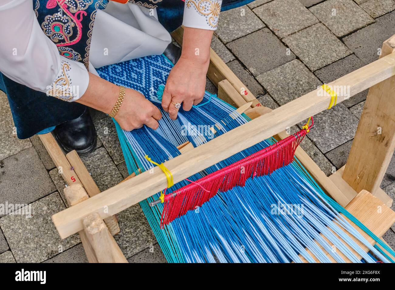 Woman's hands weaving long rug from wool threads with traditional Asian pattern on hand-held wooden loom at crafts fair, Almaty, Kazakhstan. Stock Photo