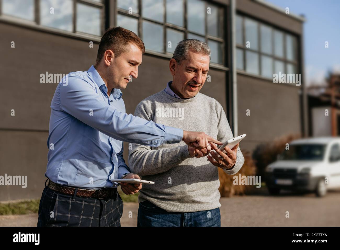 Two men are standing outside a building on a sunny day. A younger man in a blue shirt and plaid pants is pointing at a tablet in the older man's hand. Stock Photo