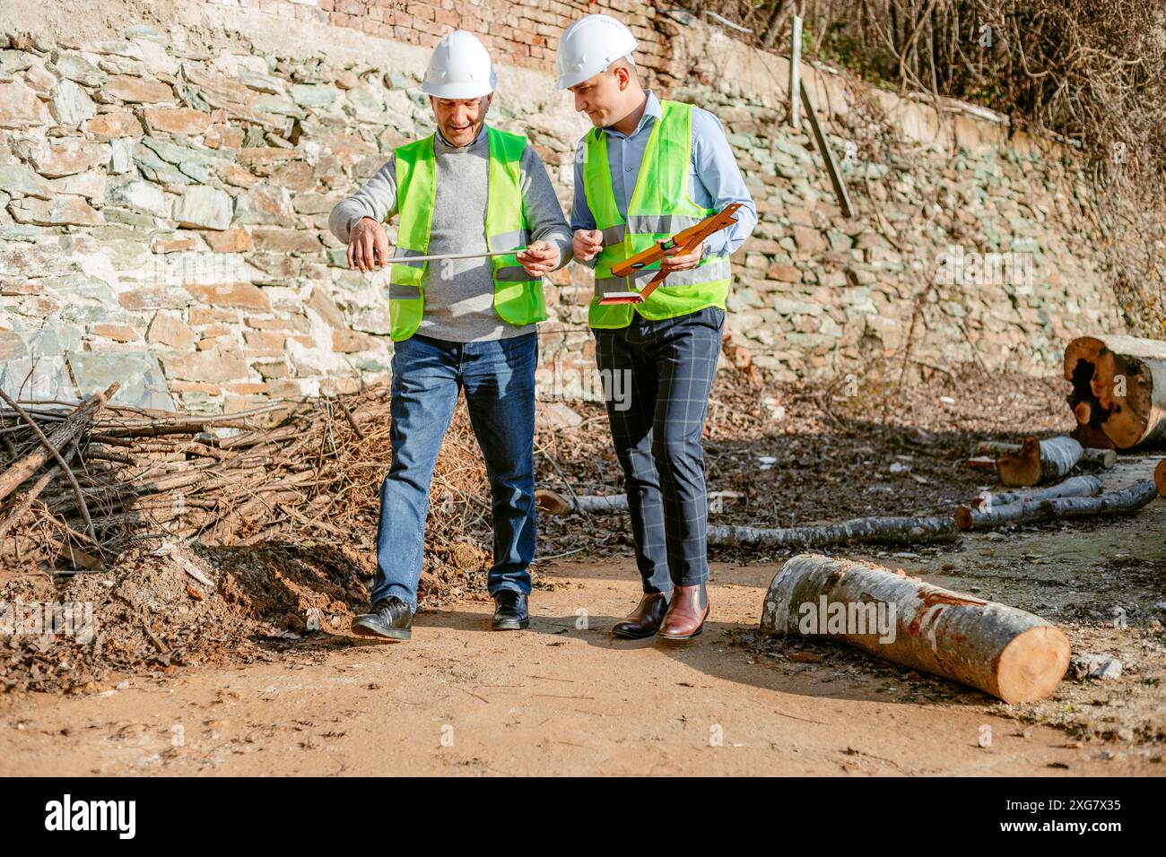Two construction workers in hard hats and safety vests walk past a pile of logs and a stone wall on a dirt path. They are working on a project at a co Stock Photo