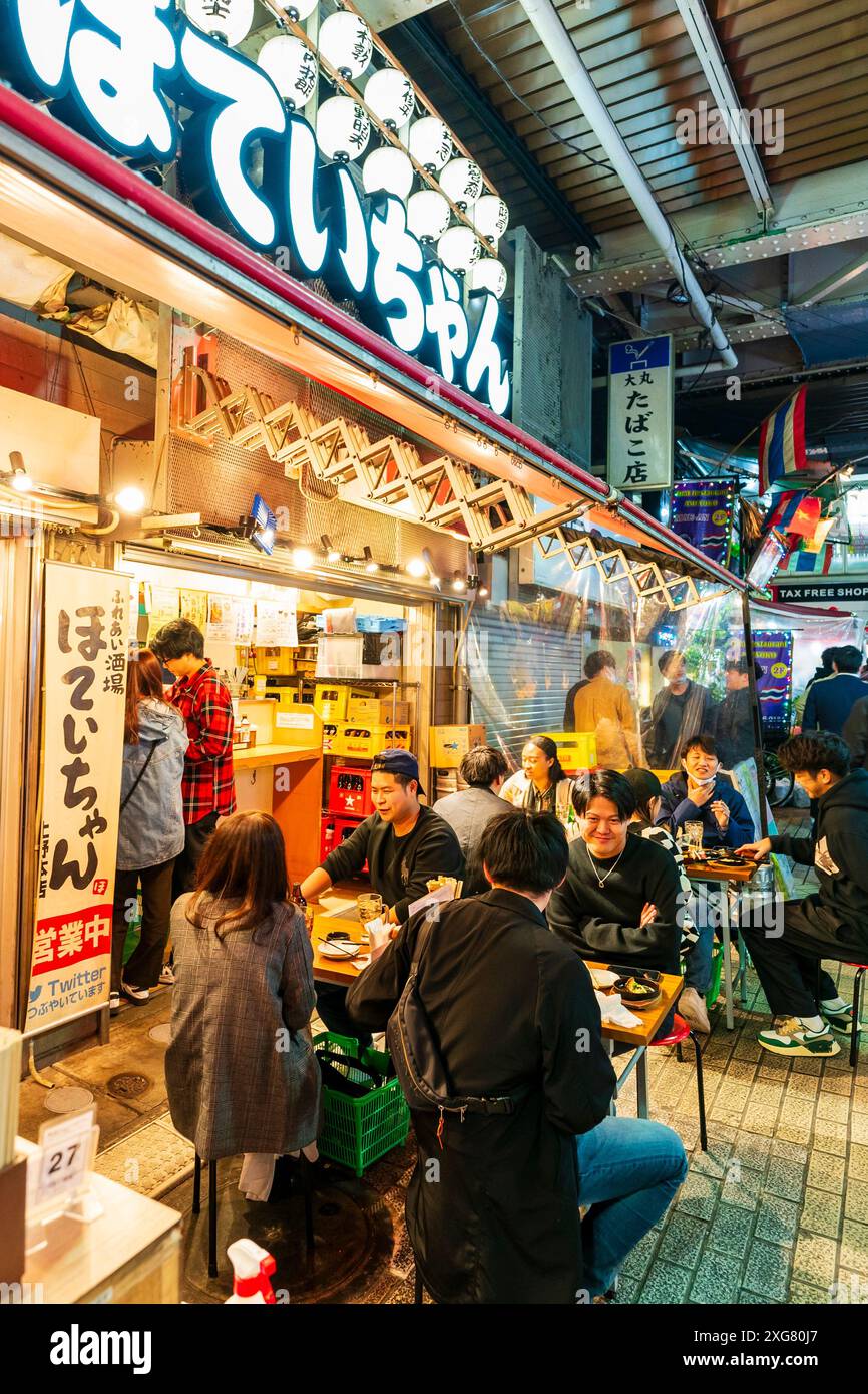 Diners sitting at tables outside a popular Japanese restaurant in the evening with people walking by, in the busy Ueno Ameyoko district of Tokyo. Stock Photo