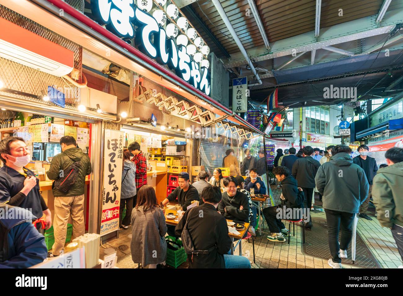 Diners sitting at tables outside a popular Japanese restaurant in the evening with people walking by, in the busy Ueno Ameyoko district of Tokyo. Stock Photo