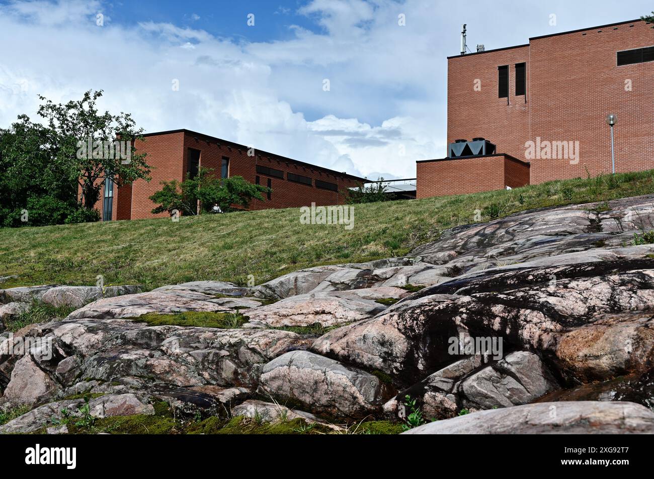 Image of two brick buildings sitting atop a rocky hill with a grassy slope, set against a backdrop of a blue sky with some clouds visible overhead Stock Photo
