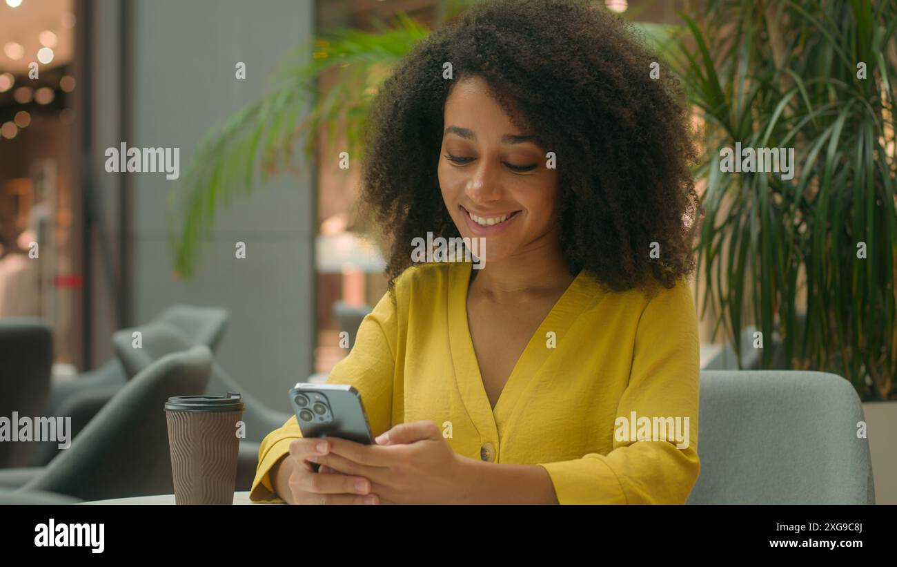African American woman in cafe drink hot tea coffee using smartphone mobile phone telephone chatting shopping mall internet ordering online texting Stock Photo