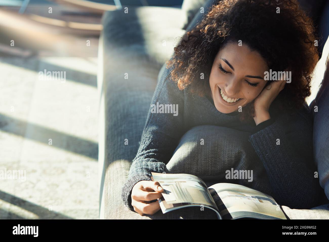 Young woman with curly hair relaxes on her couch, reading a magazine in ...