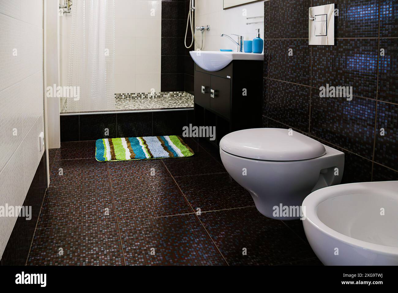 Interior of toilet with shower cubicle,floor and wall with brown tiles,sink,mirror,light fixture,floor mat,shower cubicle,curtains. Stock Photo