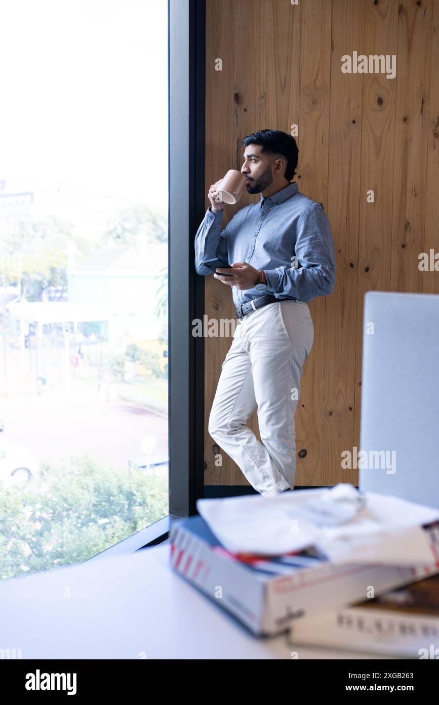 Drinking coffee and using smartphone, businessman standing by office window Stock Photo