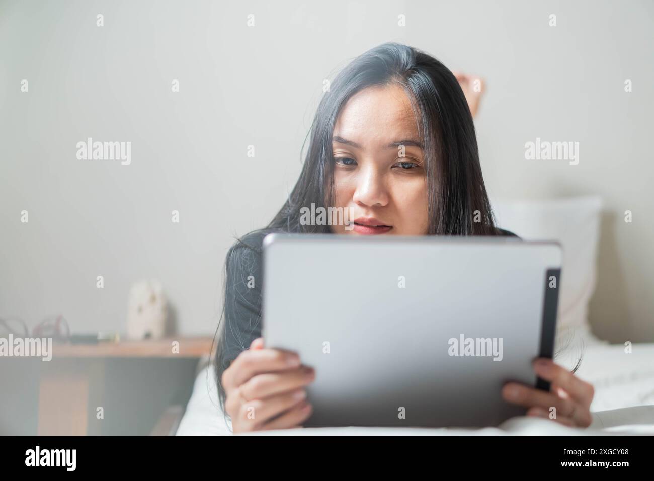 A woman lying on a bed and using a tablet. She appears to be focused on the screen. The background is minimalistic with a small table and some objects Stock Photo