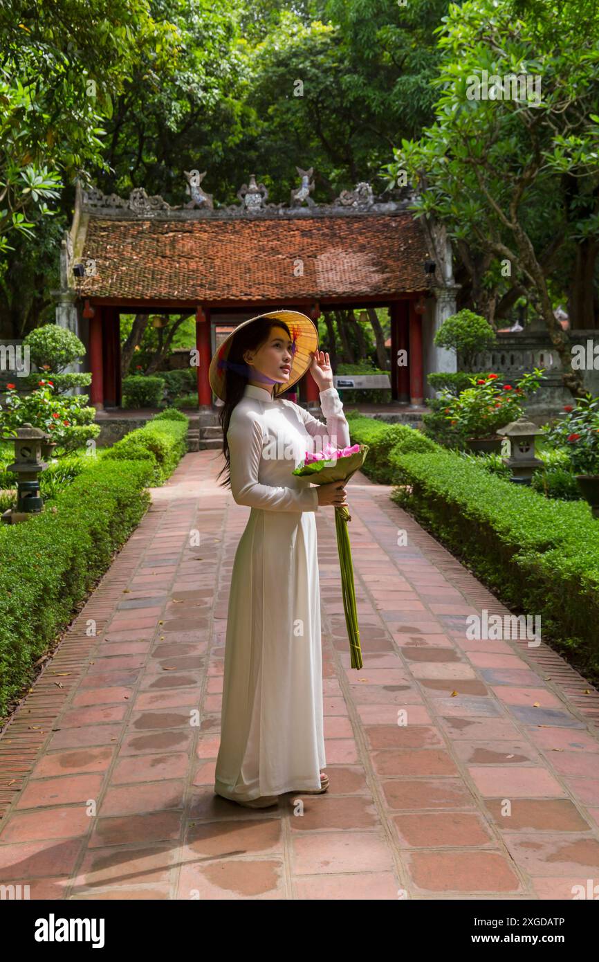 Pretty Vietnamese young woman wearing traditional Ao Dai tunic holding bunch of lotus flowers at Temple of Literature, Hanoi, North Vietnam, Asia Stock Photo