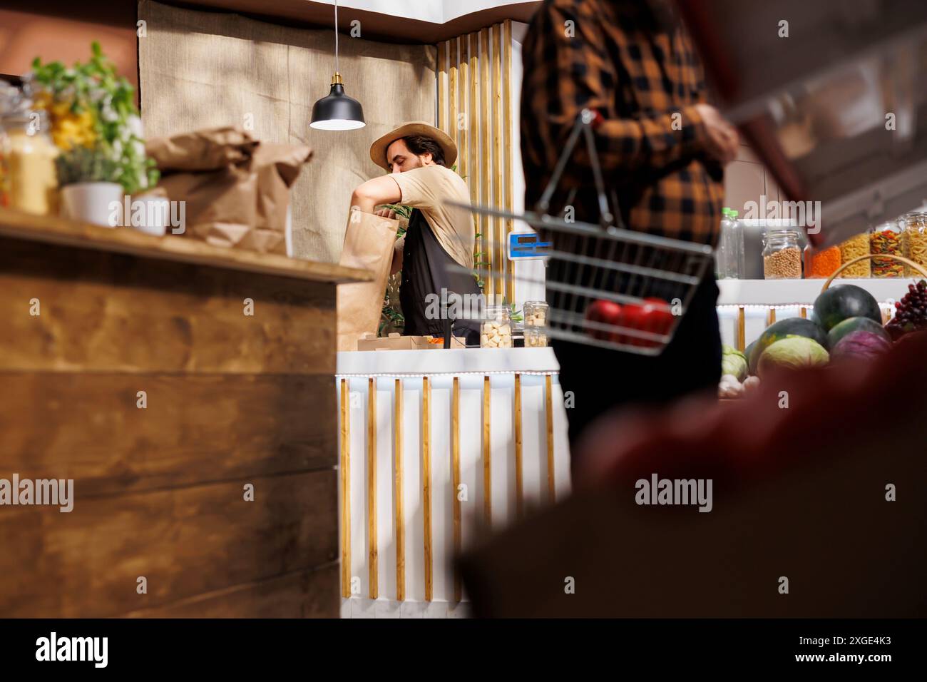 Zero waste store vendor at checkout counter bagging organic bio vegetables grown locally, while customer holding shopping basket looks for pesticides free healthy groceries Stock Photo