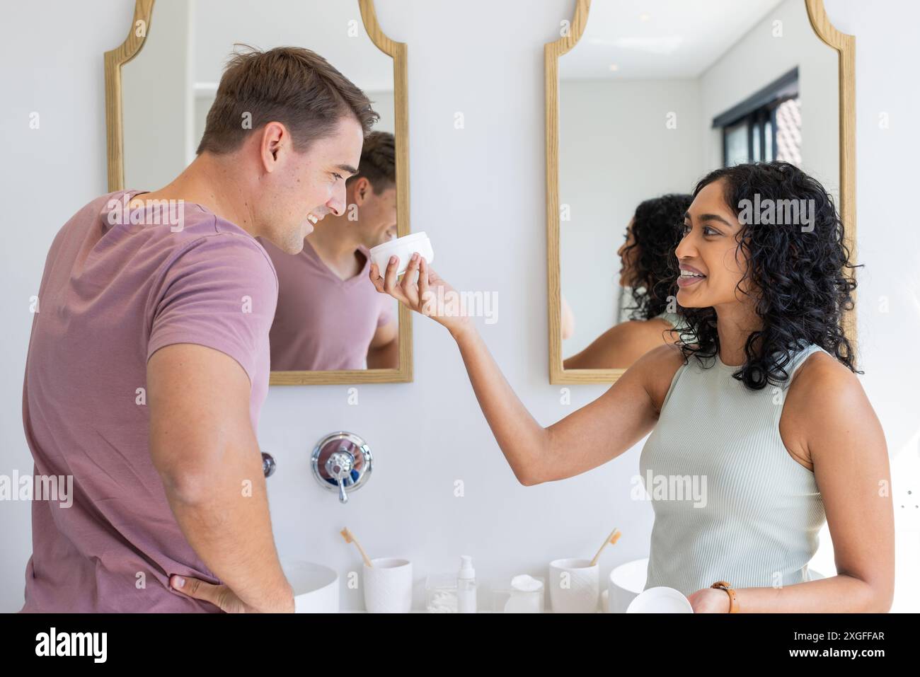 Smiling couple in bathroom, woman playfully holding skincare product to man's face Stock Photo