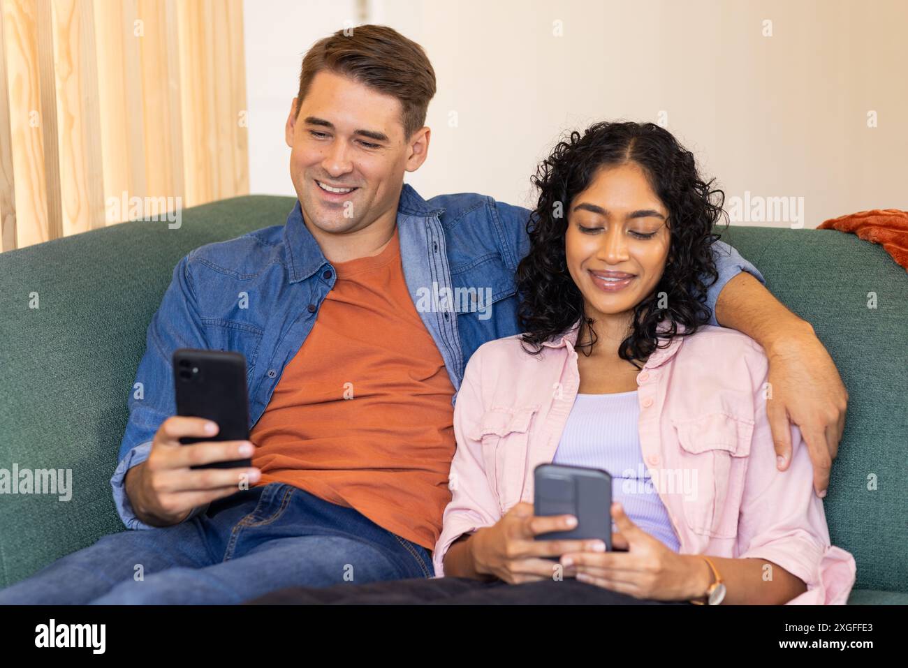 Young couple sitting on couch, using smartphones and smiling together Stock Photo