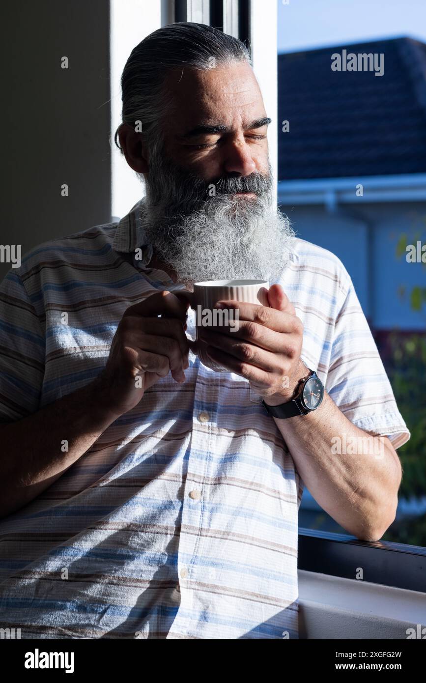 Holding coffee cup, senior man with beard enjoying morning sunlight by window Stock Photo