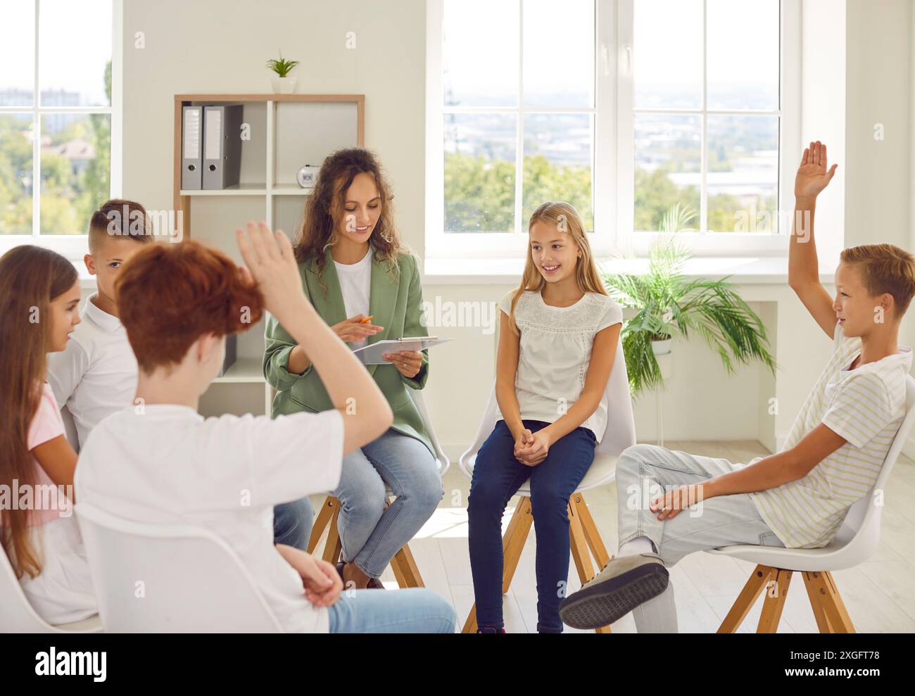 Group of school children sitting in a circle with psychologist and raising hands up. Stock Photo