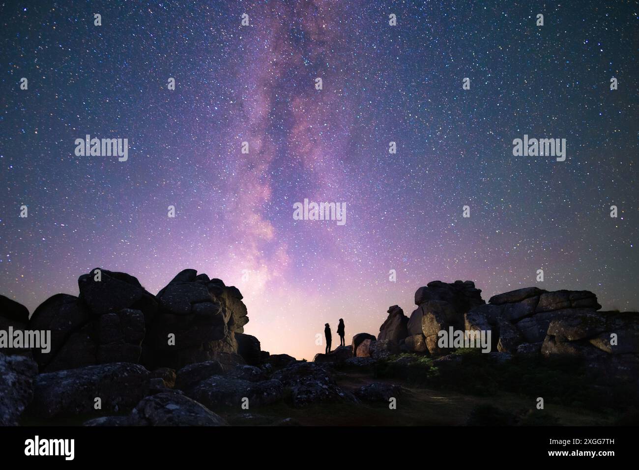 Two figures in silhouette standing below the Milky Way over a rocky Dartmoor National Park landscape at night Stock Photo