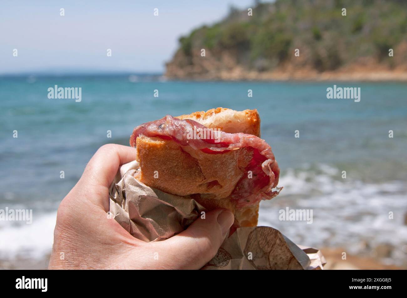 traveler eating a sandwich (panino) wrapped in paper in front of the sea, Tuscany, Italy Stock Photo