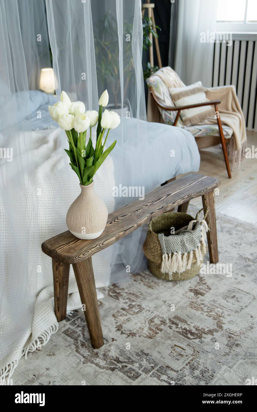 Wooden bench with a vase of white tulips. Accompanied by a cozy bed with blue linens, sheer curtains, a wicker basket, and a patterned rug, creating a Stock Photo