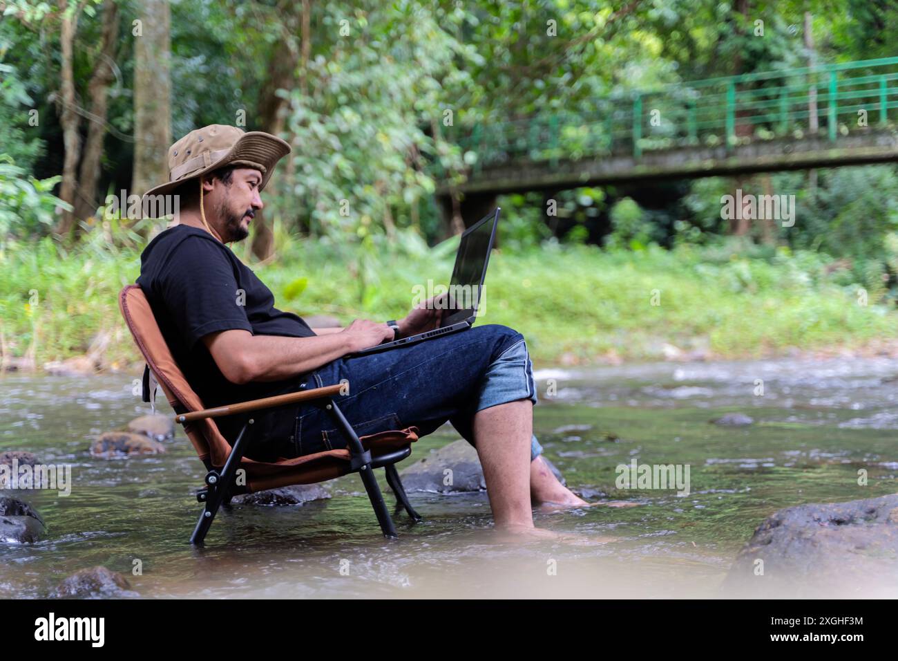 Handsome bearded Asian man of working age sitting on camping chair and working on laptop near stream in middle of park in outdoor forest alone happily Stock Photo