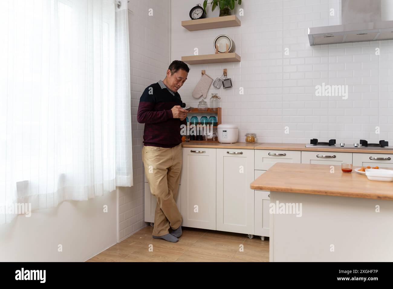 Happy middle aged old retired man in eyewear using cellphone, checking recipe online for tasty food, setting timer preparing meal alone in modern kitc Stock Photo