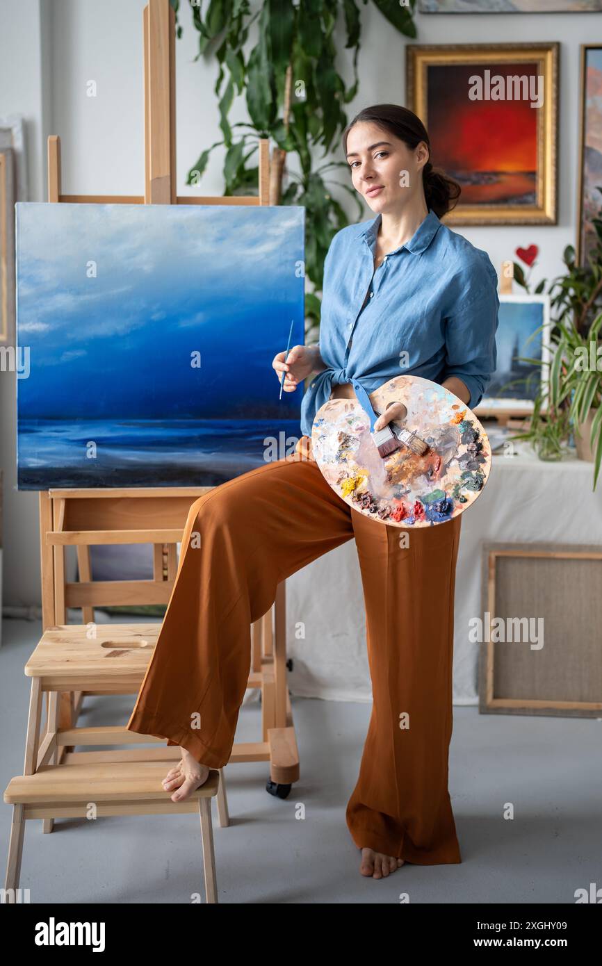 Confident artist with colour palette looks at camera stands near easel with painting in art studio. Stock Photo