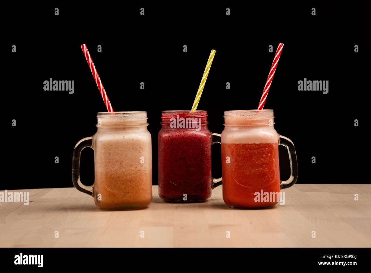 The three mason jar glasses filled with different smoothies and straws on a wooden table against black background Stock Photo