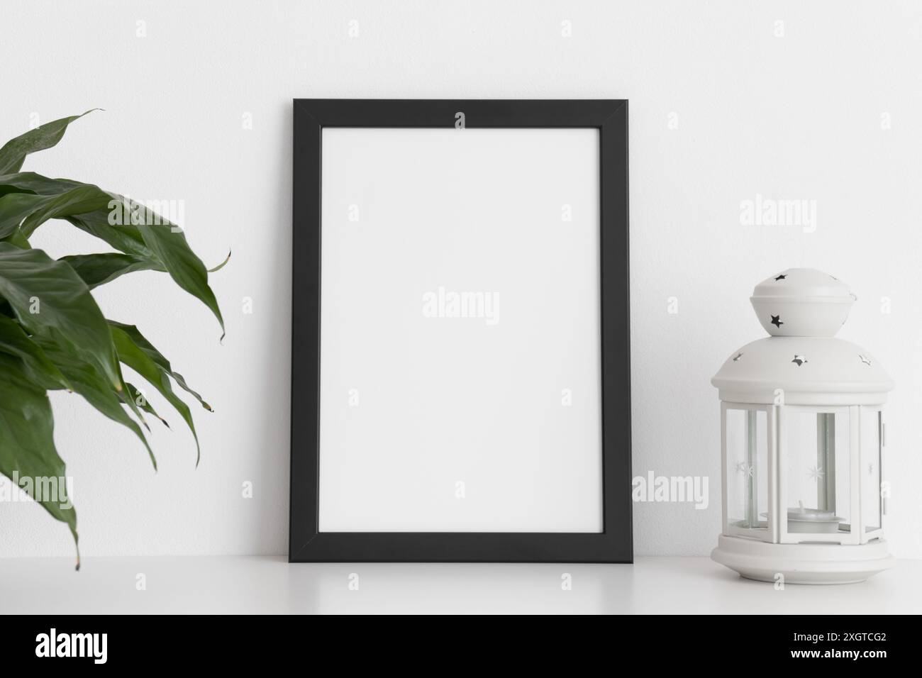 Black frame mockup with a spathiphyllum and candle holder on a white table. Portrait orientation. Stock Photo