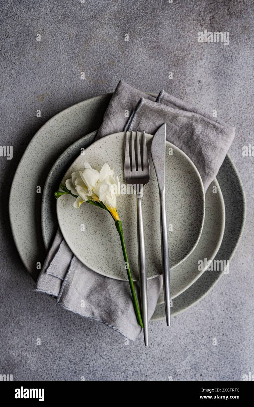 This image showcases a sophisticated table setting featuring a fresh freesia flower on speckled ceramic plates, complemented by sleek silverware and a Stock Photo