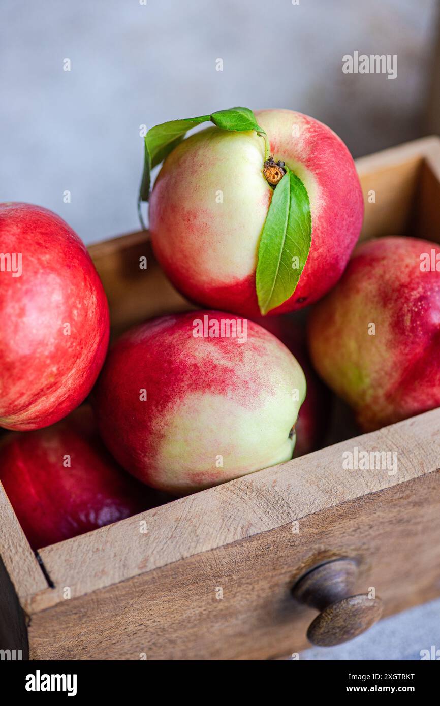 A close-up image showcasing fresh, ripe nectarines with vibrant red hues, nestled in a quaint wooden crate. Each nectarine features a tender green lea Stock Photo