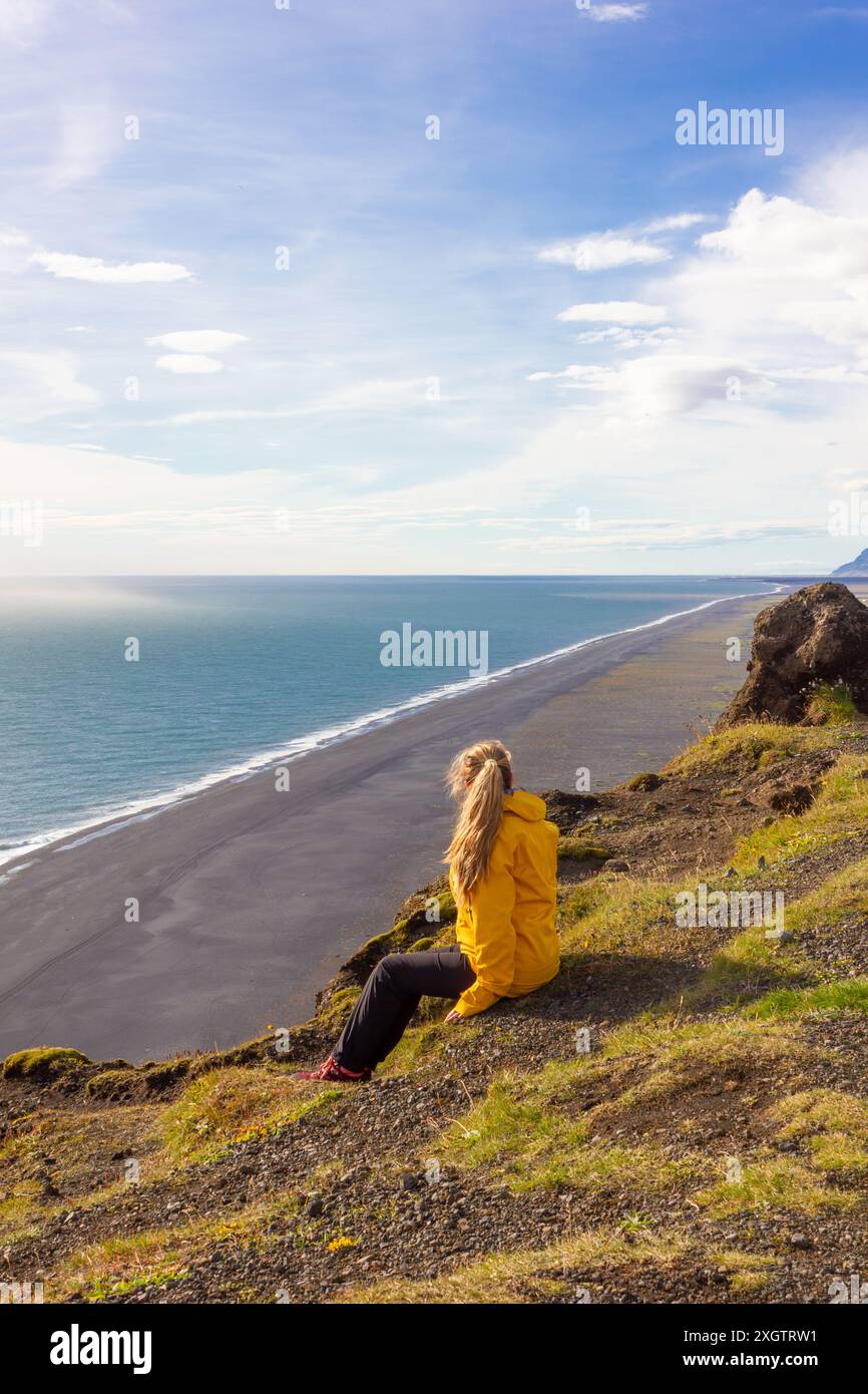 A contemplative woman sits on a cliff in a bright yellow jacket, observing the vast, tranquil black sand beach and glistening ocean under a wide blue Stock Photo
