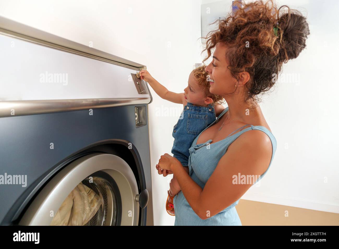 Toddler inserting a coin into the slot of the washing machine. Hispanic woman putting coin in washer machine for operated. Self-service laundry facili Stock Photo