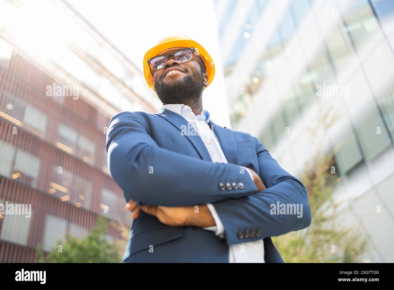 African American businessman in blue suit and yellow hard hat, standing outdoors with arms crossed and looking away confidently. Stock Photo