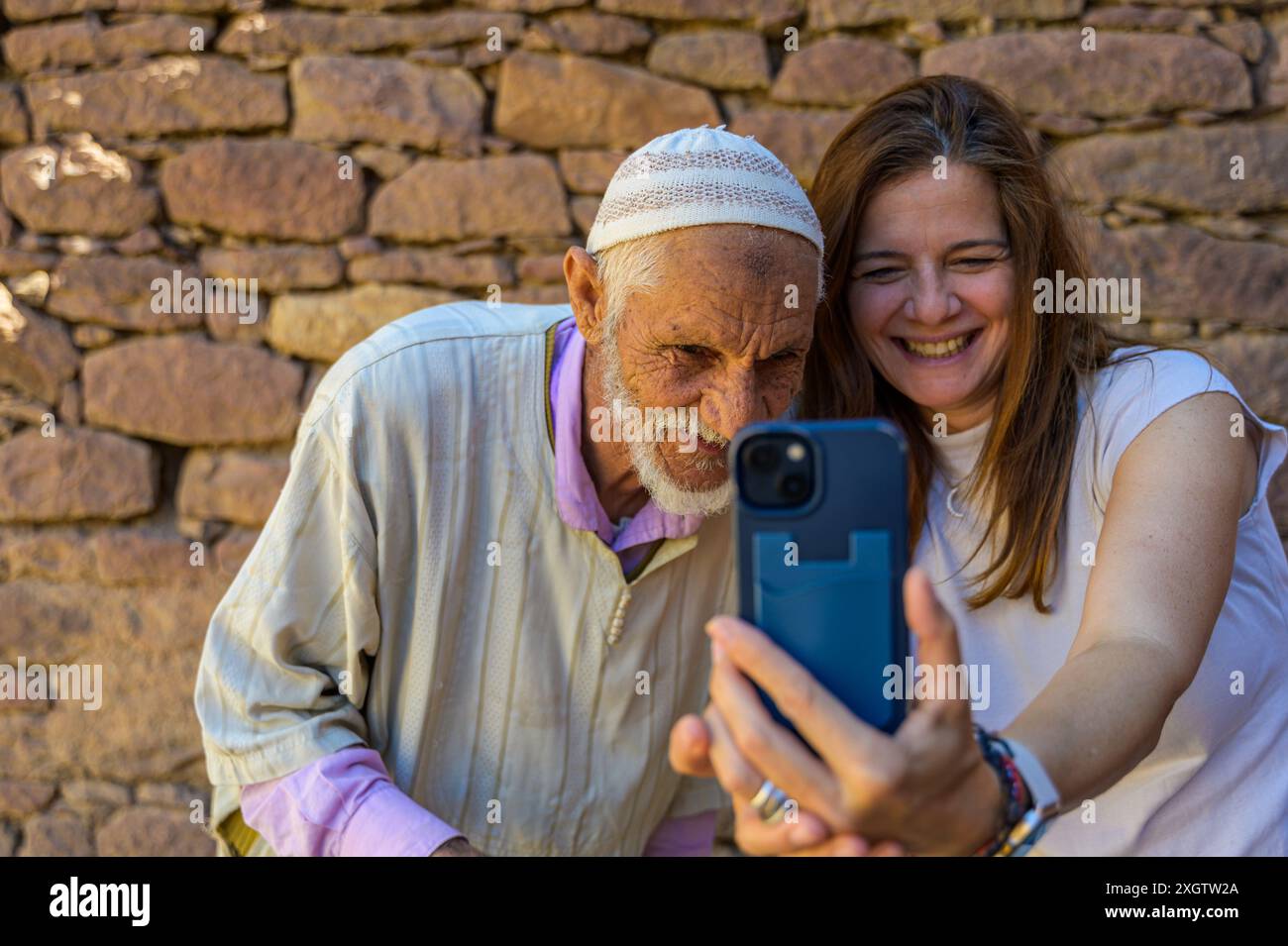 In Morocco, a smiling young woman shares a moment with an elderly man by taking a selfie together against a stone wall background The joy and intergen Stock Photo