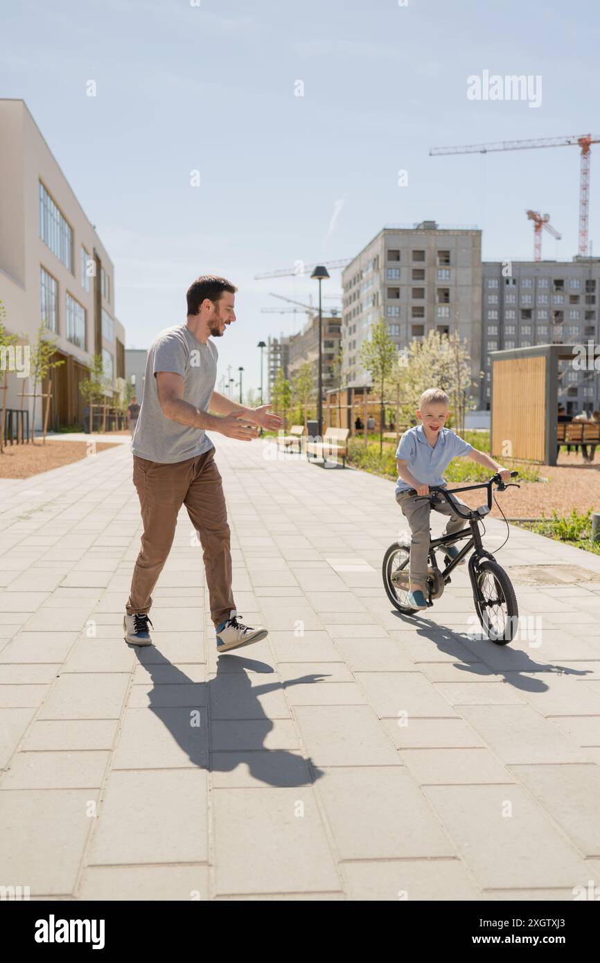 A joyful scene in an urban park where a father, looking at his son, is teaching him how to ride a bicycle. The son, looking away towards the path and Stock Photo