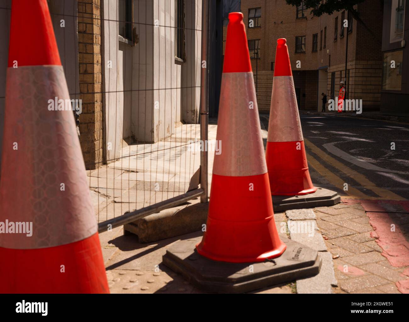 A construction site worker wearing luminous orange protective clothing with three traffic cones in the foreground at the side of a road Stock Photo