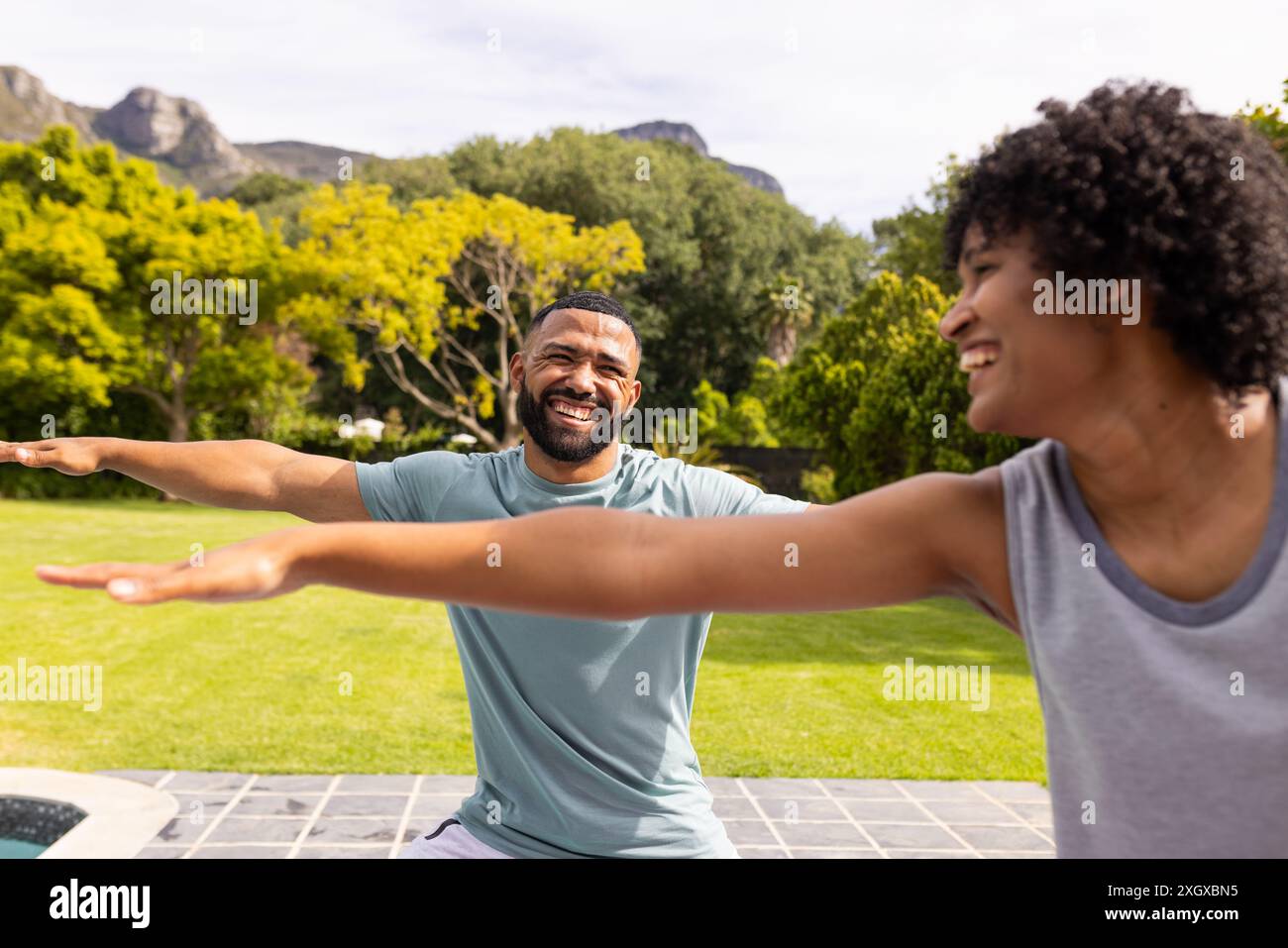 Biracial couple enjoys a fun moment outdoors doing yoga. They're sharing laughter while stretching in yoga poses in a sunny garden. Stock Photo