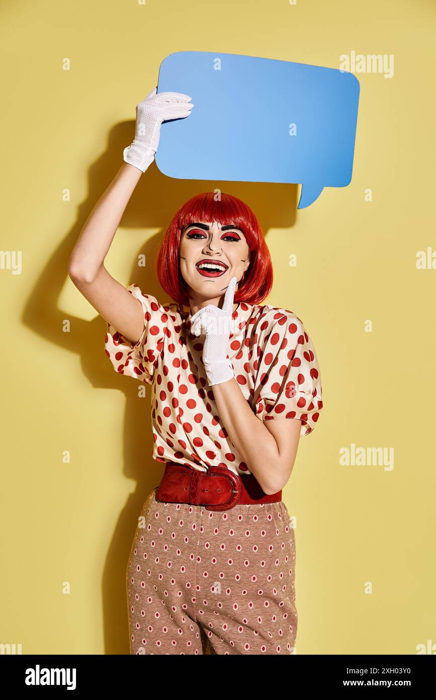 A creative redhead woman in polka dot blouse holding a speech bubble above her head on a yellow background. Stock Photo