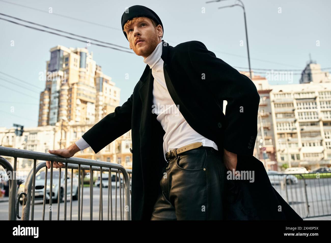 Young red-haired man leaning on rail, wearing coat and hat, exuding debonair style. Stock Photo