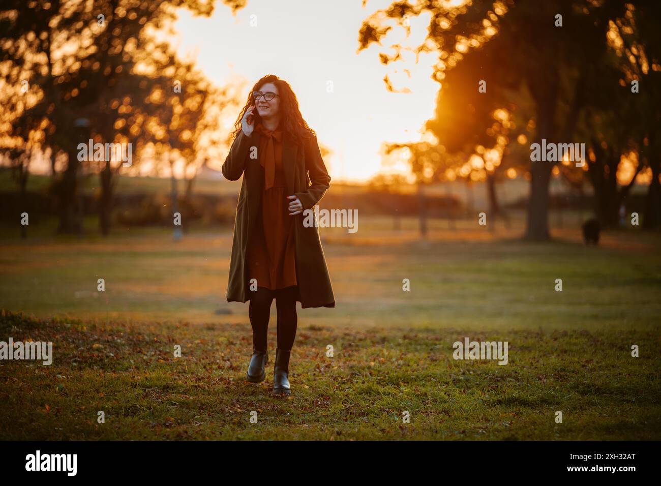 A woman in a coat and glasses walks through a park while speaking on her cell phone. The sun is setting, casting a warm glow over the scene. Stock Photo