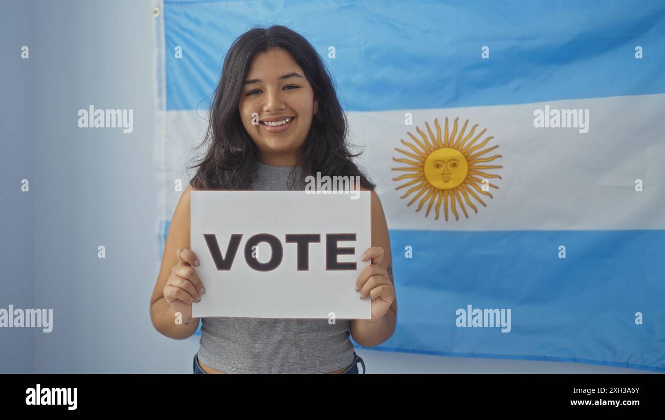 A young, smiling woman holds a vote sign with an argentine flag in the background, emphasizing an electoral setting indoors. Stock Photo