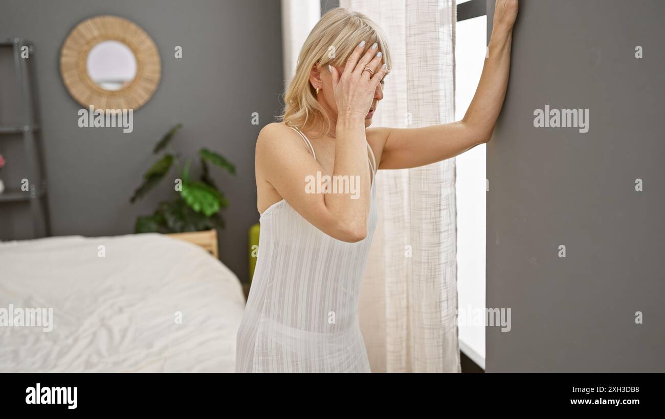 A young, attractive blonde woman in a bedroom setting, touching her head as if distressed. Stock Photo