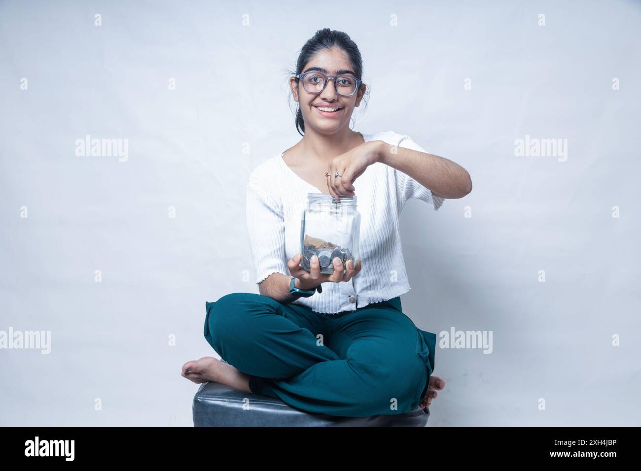 happy Indian school girl putting coin in glass jar while sitting on sofa. education, saving
