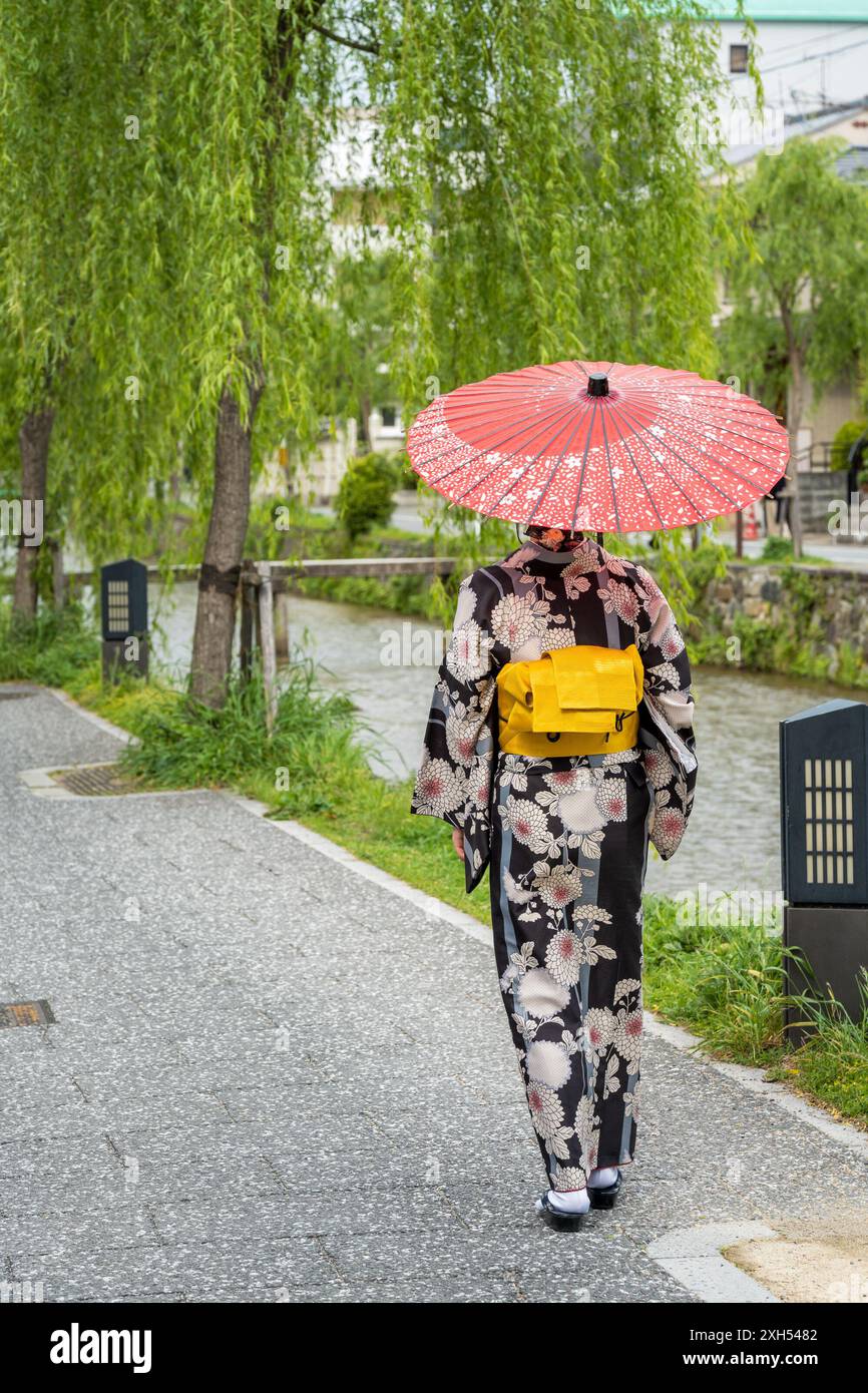 Japanese woman in vibrant traditional kimono holding red umbrella on Kyoto street during rain, demonstrating the aesthetic beauty of kimono rental during wet weather conditions