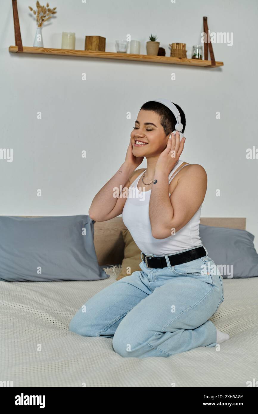 Young woman with short hair, dressed in casual wear, sits on a bed ...
