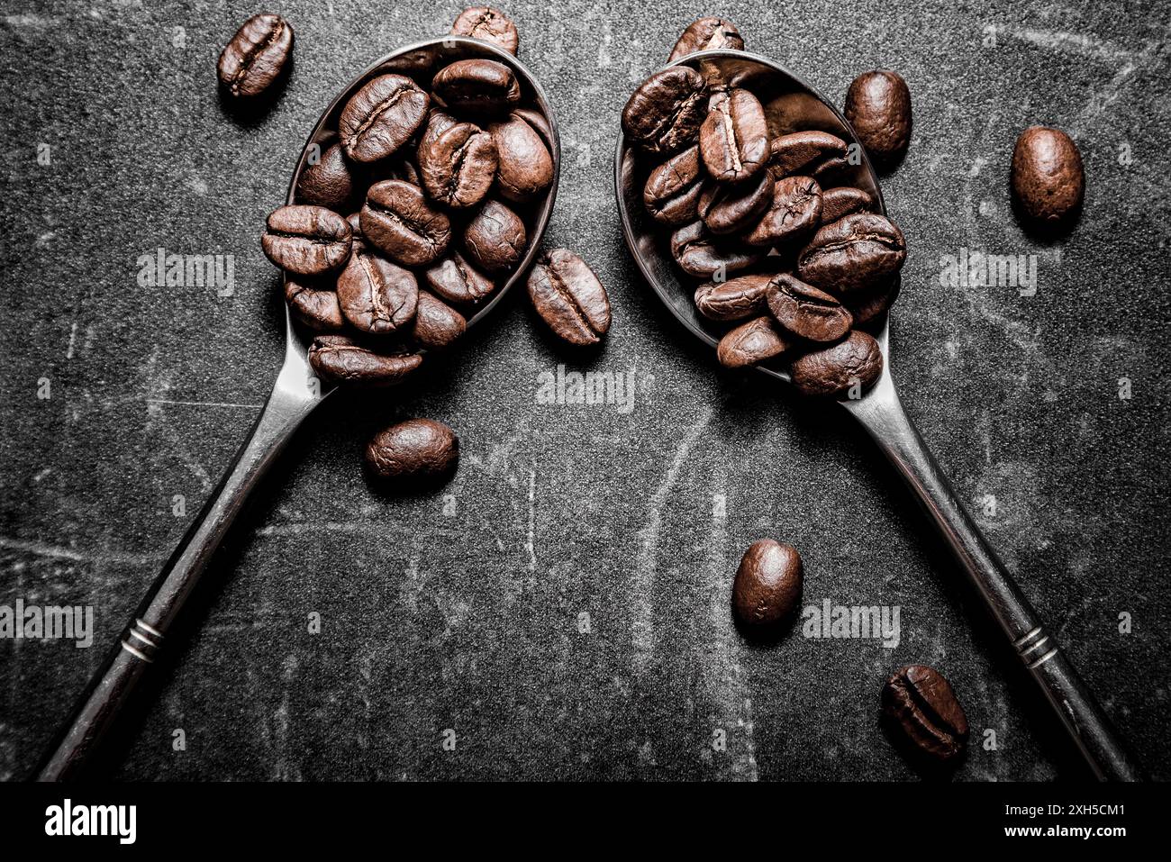 Two silver spoons are filled with glossy brown coffee beans against a dark, textured background. Several loose coffee beans are scattered around Stock Photo