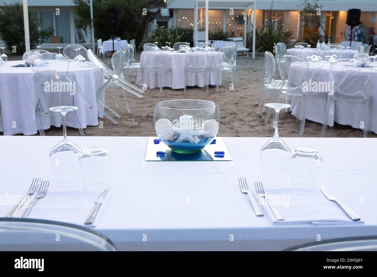 Round tables with white tablecloths and transparent chairs set up for an event on the beach Stock Photo