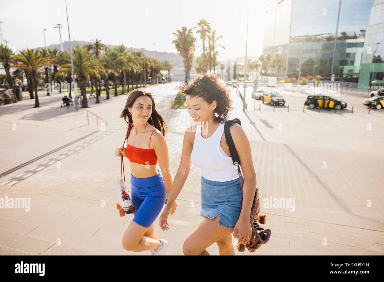 Two young women are walking up stairs together, smiling and chatting, each holding a pair of roller skates, ready for a day of skating fun. Stock Photo