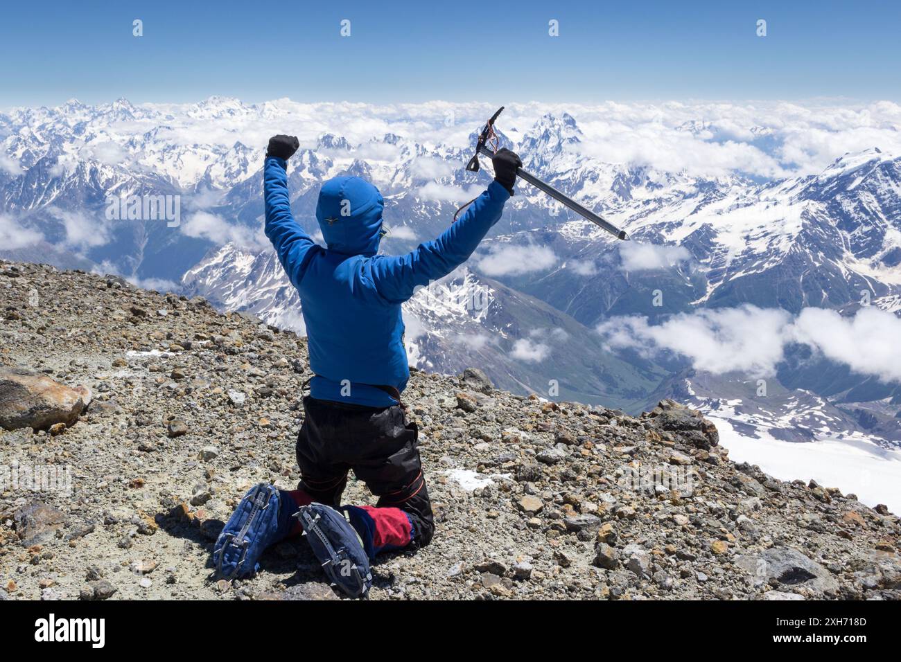 Mountaineer man in crampons and blue jacket on summit of mountain sitting on his knees holding ice axe and throws hands up Stock Photo