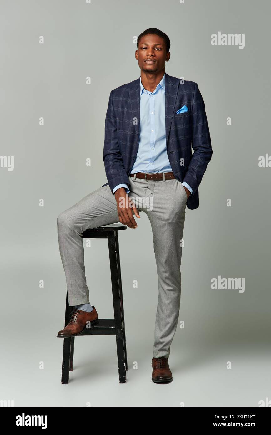 Handsome African American man in a stylish checkered blazer and classic  suit, sitting on a sleek stool against a grey background Stock Photo - Alamy